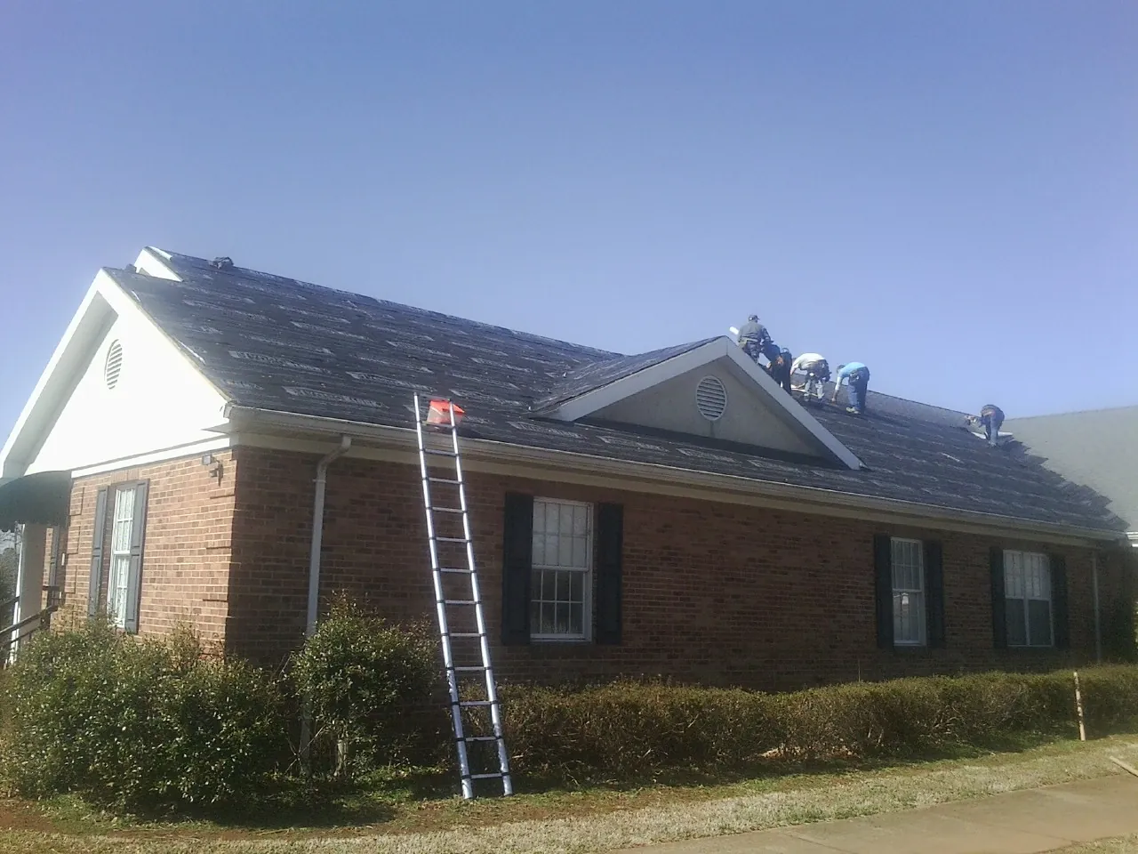 Crew tearing shingles off a brick ranch roof with a ladder set — Metal Roof Repair work by our crew in Elliott